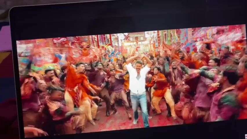 The video displays a close-up of a laptop screen, showing a scene from a movie or music video. The scene is a vibrant, colorful outdoor celebration with a large crowd of people dancing. A man in a white shirt and blue jeans is the central figure, dancing and interacting with the crowd. The crowd is dressed in colorful clothing, and there are decorations and structures visible in the background, suggesting a festival or parade. The overall atmosphere is energetic and joyful, with confetti or petals scattered on the ground.
