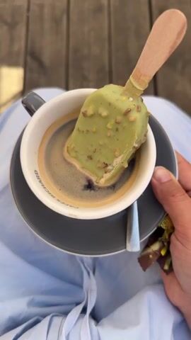 A person's hands hold a dark gray mug and saucer filled with dark coffee. A pistachio-flavored ice cream bar with nuts on the coating is submerged halfway into the coffee. The ice cream bar has a light-colored wooden stick. The person is wearing a light blue garment. The scene appears to be outdoors on a wooden deck, with some blurred background elements that suggest outdoor seating. A spoon rests on the saucer.