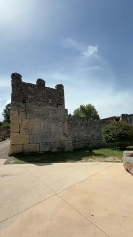 The video captures an outdoor scene at the Conjunt Monumental d'Olèrdola, located in Vilafranca del Penedès, Spain, on a bright, sunny day. The camera begins by facing a tall, weathered stone wall, part of ancient ruins, with a clear blue sky above. The wall features a battlements-like top and is constructed from large, light-colored blocks.

As the camera pans to the left, more of the historical site is revealed. A large, mature tree with lush green foliage comes into view in the foreground, casting shadows on the paved pathway. Beyond the tree and pathway, additional sections of the stone walls and ruins are visible, set against grassy areas. The panoramic view emphasizes the expansive nature of the archaeological site.

A prominent red informational sign is positioned at the base of the large tree. The sign clearly displays the text "Conjunt Monumental d'Olèrdola," indicating the name of the monument or archaeological complex. Smaller text and institutional logos are also visible on the sign. The scene conveys a sense of history and open-air exploration, typical of an ancient monument. No people are visible in the video.