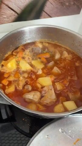 A close-up, overhead shot shows a metal pot filled with a simmering stew on a stovetop. The stew contains chunks of meat, potatoes, carrots, and peas in a rich, reddish-orange broth. A wooden spoon is used to stir the contents of the pot. The background is slightly out of focus, showing a wooden-patterned surface. Another metal lid is visible in the foreground.