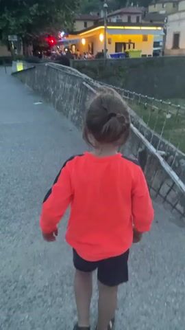 This video captures a young girl walking away from the camera on a paved pathway. She is wearing a bright orange sweatshirt with dark sleeves and dark shorts. Her hair is pulled back in a bun. The pathway is bordered by a stone wall with a metal railing. Beyond the wall, a dried-up riverbed is visible, with patches of grass.

In the background, across the riverbed, is a village scene. Buildings with lit windows suggest that it is evening. One building, with yellow lighting, appears to be a restaurant or cafe with people seated outside. Further back, more buildings line the side of a hill, and mountains are visible in the distance. The sky is cloudy, indicating it might be dusk or a cloudy day. A red traffic light is visible on a pole near the illuminated building. The overall mood is peaceful and casual, possibly a family walk in a small Italian town like Pescia.
