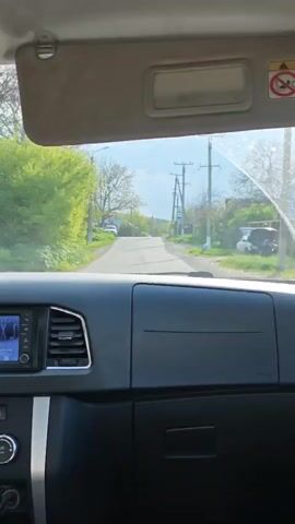 From inside a vehicle, the camera shows a view through the windshield as the vehicle travels on a paved road through a village or suburban neighborhood. Trees with green foliage line the sides of the road, and telephone poles are visible. Houses, some with visible rooftops, dot the landscape. The sky appears partly cloudy. The interior of the vehicle is visible, featuring a dashboard with a built-in screen displaying what appears to be navigation information or media controls. A sun visor is partially extended on the driver's side. The general atmosphere suggests a daytime drive in a peaceful, residential area. The windshield is partially wet.