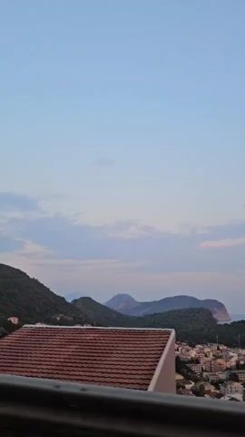 The video shows a slow pan over a coastal town nestled among hills. In the foreground, a red-tiled roof of a building is visible. The middle ground features houses with red roofs, and further back, mountains rise against a pale blue sky with wispy clouds. The overall impression is one of a serene, possibly European, coastal landscape during either sunrise or sunset.