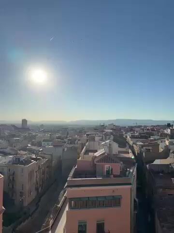 The video presents a high-angle, panoramic view of a sunny coastal city, identified as Tarragona, Spain. The camera slowly pans across the urban landscape, revealing various elements.

The initial shots showcase a dense collection of buildings, primarily residential and commercial, with diverse architectural styles, many featuring light-colored facades (white, beige, pink) and flat or low-pitched roofs. In the background, distant mountains are visible under a clear, bright blue sky, with a strong sun glare dominating the upper left portion of the frame. A faint aircraft contrail can be seen high in the sky.

As the camera continues to pan to the left, a significant body of water, the Mediterranean Sea, comes into view, stretching towards the horizon. The coastal perspective reveals a large, modern white hotel building nestled among other city structures. Below, a busy road with visible car traffic winds through the city, featuring a prominent roundabout. Ancient, weathered stone walls and ruins, characteristic of Tarragona's Roman heritage (likely remnants of the Roman city walls or amphitheater), are also clearly visible, contrasting with the newer developments.

The overall scene depicts a vibrant city under a brilliant, clear sky. The time of day appears to be late morning or early afternoon, given the sun's position and intensity. The weather is ideal, with no clouds and excellent visibility. No people are distinctly visible, and there is no discernible text within the video. The atmosphere is serene and inviting, highlighting the city's historical depth and picturesque coastal setting.