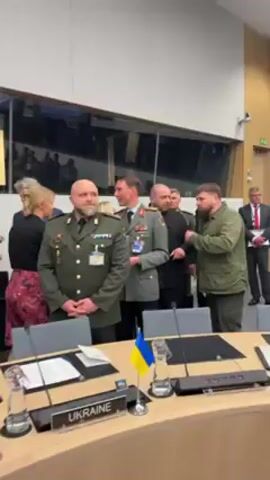 Here is a concise description of the video:

The video captures a group of people in what appears to be a conference setting. Several individuals are wearing military uniforms, including one with a German flag insignia. A Ukrainian flag is visible on a table in the foreground. The people in the scene appear to be engaged in conversation, with some shaking hands and smiling. The setting is a well-lit room with a view of an area filled with tables and seated individuals in the background. The overall atmosphere seems cordial and professional.