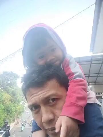 A low-angle, close-up, selfie-style video captures a man giving a child a piggyback ride. The man's face, partially obscured by the child, is in the foreground. The child, wearing a pink hooded jacket, sits on the man's shoulders, looking towards the camera. The background shows a street scene with trees and foliage on the left, and a building with a corrugated roof on the right. A few motorcycles are visible on the road. The lighting suggests it is daytime, possibly overcast.
