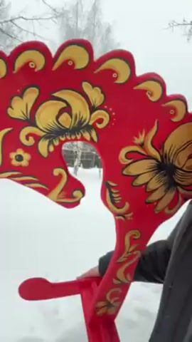 A person with long light-brown hair and a red beard, wearing a black puffer jacket, winks at the camera while holding a red wooden rocking horse decorated with yellow and black floral designs. The person is standing in a snowy landscape with trees in the background.