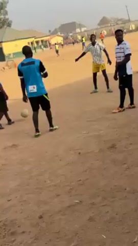 The video shows a group of young men playing soccer on a dusty field. Several men are wearing soccer jerseys and shorts. One man in a light blue jersey with black shorts appears to be instructing or coaching others. He's gesturing with his hand, and one man is kicking the soccer ball in his direction. Other men are standing around watching or participating in the drill. In the background, there are more people on the field, and a building with a green and yellow roof is visible. The environment appears to be an outdoor soccer field with dry, dusty ground.