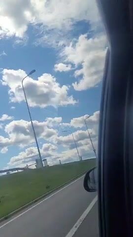 The video shows a view from inside a vehicle, looking out the side window. The view is of a road, a grassy embankment with some structures, streetlights, and a blue sky with white, fluffy clouds. A large white truck with a tanker trailer passes by on the road. The sun is shining, and it appears to be daytime.