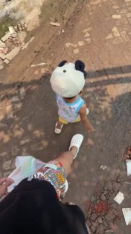 A toddler walks away from the camera on a dirt and brick path. The toddler wears a white panda-themed hat, a sleeveless shirt with a colorful design, and yellow shorts. The toddler's legs are blurry, indicating movement. The camera is positioned low, looking up at the toddler. The foreground shows the legs and feet of an adult wearing patterned shorts and white clogs, with a white plastic bag in their left hand. Sunlight casts shadows on the ground, suggesting it is daytime. The environment appears to be outdoors with a rough, unpaved surface.