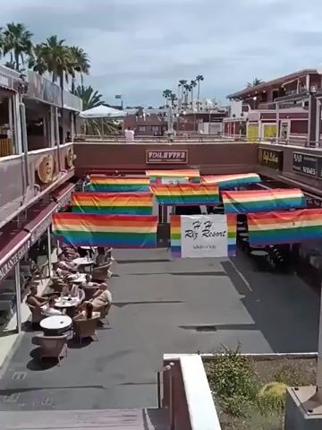 The video shows a view from a balcony overlooking a commercial area. Below, several rainbow flags are hung, and a sign reads "H. H. Riz Resort Adults Only". People are seated at tables in what appears to be an outdoor dining area. Signs for businesses like "Eiffel Music Wines" and "Foot on Mars" are visible. The environment includes palm trees and a partially cloudy sky. The overall impression is that of a vacation or tourist destination.