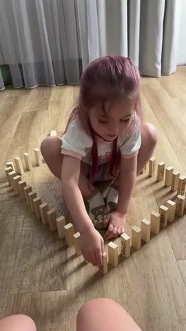 A young girl with purple hair, wearing a white t-shirt and purple shorts, is sitting on a wooden floor surrounded by dominoes arranged in a heart shape. She is playing with the dominoes, pushing one over to start a chain reaction. She laughs as the dominoes fall. The background shows a window with white curtains.