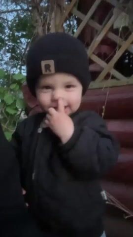 A young child, around toddler age, is the focal point of the video. The child is wearing a black beanie with an "R" patch and a black puffer jacket. The child is smiling and pointing directly at the camera.  The child can be heard saying "Daddy." The background is somewhat blurred but appears to be an outdoor setting with a wooden structure and some greenery. The lighting suggests it is dusk or dawn.