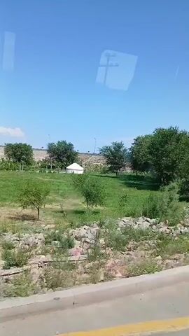 The video shows a view from a moving vehicle looking out a window. The landscape is predominantly green with trees and grass. In the distance, there is a white yurt-like structure and a large metallic sculpture that appears to be a statue of a person or animal. The sky is clear and blue. The road or surface the vehicle is on is visible in the foreground, with a yellow line painted on it. The scene suggests a park or an outdoor recreational area.
