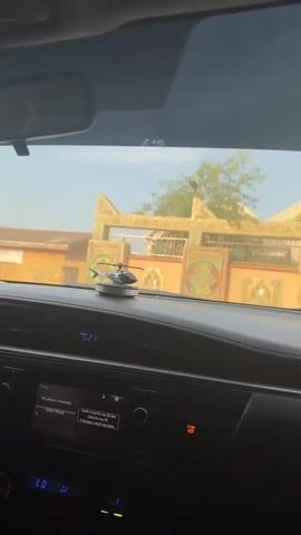 A car is driving down a street with yellow or tan buildings visible through the windshield. A small helicopter ornament is on the dashboard. The car's information screen says "No phone connected" and "Unable to read the map SD data." The hazard lights button is illuminated. The car horn honks.