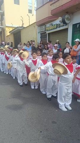 A line of children is walking down the middle of a street. All of the boys are wearing white pants and jackets, with red bandanas around their necks and straw hats in their hands. The girls are wearing white and red dresses. The children are walking in a straight line, holding their hats, and looking towards the camera. Spectators are standing on the sidewalks, watching the procession. The street is paved, and the buildings are a mix of residential and commercial. A store called "Mr.Gold" is visible in the background. There are trees and parked cars in the background as well.  The scene appears to be a street parade or cultural celebration.