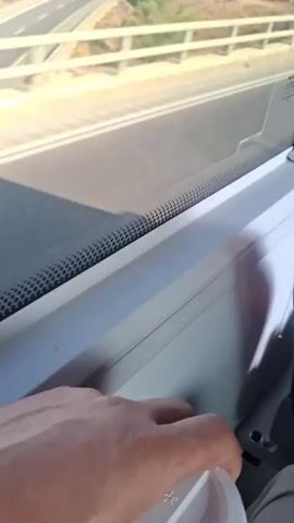 The video is shot from inside a vehicle, likely a bus or train, looking out through a window. The perspective is slightly tilted, suggesting movement. Outside the window, a highway is visible, with cars traveling in the distance. The landscape is dry and arid, with patches of sparse vegetation. The sky is clear and blue. Reflections on the window obscure some details, but the overall impression is of a journey through a desert-like environment.