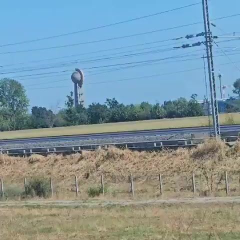 The video shows a tall, cylindrical structure with a spherical object on top, partially obscured by trees. This structure is visible against a clear blue sky, with power lines running across the frame. In the foreground, there is a train track with overhead electrical wires. The surrounding environment appears to be rural, with dry grass and vegetation visible. The camera appears to be moving, possibly from a vehicle, as the perspective shifts slightly throughout the short clip. There are no people or animals visible in the video. The structure itself is the primary subject, and its unique design suggests it might be a water tower or a monument. The overall impression is one of a passing view of a somewhat unusual landmark.