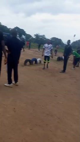 The video shows an outdoor scene on a dirt ground. Several people are present, some wearing black shirts with white lettering on the back that appears to say "POLICE" or "NPF". There are also people wearing athletic clothing in red and yellow. In the background, there are trees and some mountainous terrain. In the foreground, two individuals in black shirts and black pants with white stripes are standing, facing away from the camera. One of them appears to be holding a camera or recording device. In the middle ground, there are several car tires arranged on the ground, possibly as an obstacle course. Some individuals are interacting in the distance, appearing to engage in some form of activity or training. The overall atmosphere seems to be that of an organized event or training session taking place in an open, rural environment.