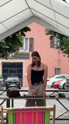 A person with long hair, wearing a black tank top, beige shorts, and large pink earrings, stands under a white tent, operating a DJ mixer and laptop. Behind them is a pink building with dark shutters and a sign that reads "JF REPARATIONS MULTIMARQUES". A black car and two other cars are parked in the background. The foreground features a striped chair. The person appears to be focused on the DJ equipment.