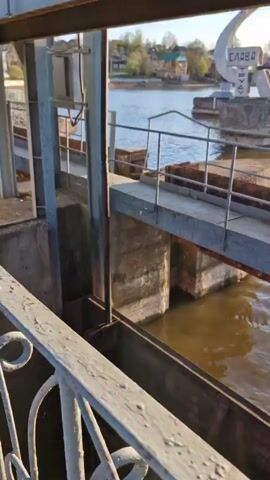 The video shows water flowing through an open dam gate. The camera is positioned above the gate, looking down into the rushing water. The water appears brown and murky, cascading into a lower level with foamy white water. Steel and concrete structures are visible around the dam. In the distance across the water, there are some houses and trees. The sky is sunny and bright. There is a monument with the word "Слава" (Glory) on it visible in the background.