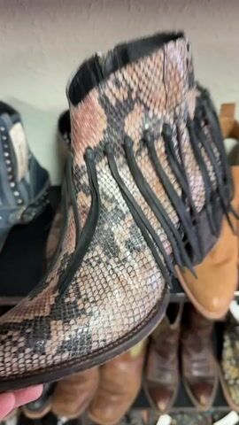 The video shows a person's hand holding and displaying several pairs of cowboy boots on shelves. The boots vary in style, color, and material. Some boots are patterned with snakeskin or leopard print, while others are solid colored in shades of brown and blue. Several boots feature decorative elements like fringe, studs, and ornate stitching. The environment appears to be a retail space or a storage area, possibly a shoe store or a personal collection, with multiple shelves filled with footwear. The lighting is somewhat dim, suggesting an indoor setting.