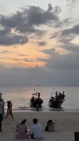 The video shows a beach scene at sunset. Two traditional longtail boats are partially submerged in the shallow water near the shore. People are scattered along the beach; a couple sits on the sand in the foreground, while others stand or walk nearby. The sky is filled with gray clouds illuminated by the fading sunlight, with a red glow visible on the horizon where the sun is setting.