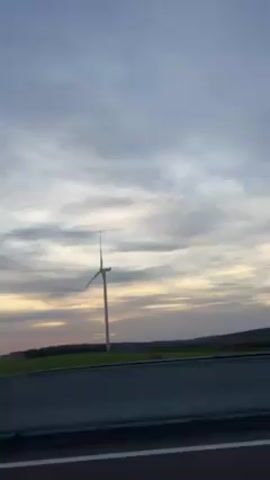 The video shows a view from a moving vehicle, likely on a highway or divided road. The sky is overcast with the sun low on the horizon, creating a soft sunset effect. Two wind turbines are visible in a field adjacent to the road. A silver car passes on the road, moving in the same direction as the camera. The scene is generally calm and rural.