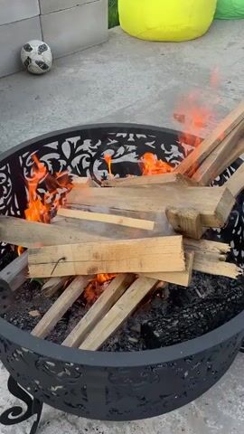 A fire burns within a decorative, circular fire pit with a lattice design along its rim. Someone adds more wooden planks to the fire. The fire pit sits on a concrete patio. A soccer ball, concrete blocks, and two beanbag chairs, one yellow and one green, are nearby.  A pile of dirt is visible in the background.
