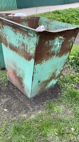 The video shows a person wearing blue and gray work gloves throwing the gloves and a red plastic object into a dumpster. The dumpster is green with patches of rust and contains several white plastic bags, a black plastic object, and a clear plastic-wrapped case of glass bottles. The dumpster is situated in a grassy area next to a paved road. A green wooden fence and a house are visible in the background.