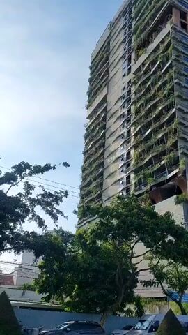 The video shows a tall, unfinished skyscraper covered in dense green foliage. The building appears to be under construction or abandoned, with visible structural elements and exposed concrete. The foliage is growing from balconies and ledges, creating a unique, almost organic facade. The sky is visible, indicating an outdoor setting. Trees and other greenery are present at the base of the building, suggesting an urban environment. Other buildings, some modern and completed, are visible in the background, contrasting with the raw state of the skyscraper. Power lines are also visible crisscrossing the frame. There are no people clearly visible or any discernible actions occurring in the video. The overall impression is one of a partially completed or neglected structure being reclaimed by nature.