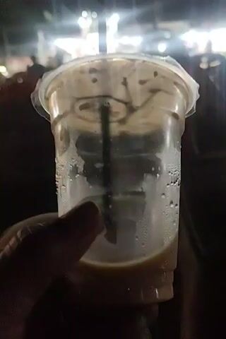 A hand is holding a mostly empty plastic cup with a straw. There is a bit of light brown liquid at the bottom of the cup, and condensation on the sides. The cup has a plastic lid. The background is blurry and dark. The scene is outdoors, possibly at night with streetlights visible.