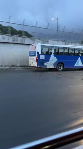 This media file captures a street scene in Quezon City, Philippines, likely during the late afternoon or early evening, given the overcast and somewhat dark sky, suggesting potential for rain or an approaching storm. The primary subjects are vehicles on a road. A blue and white bus with the license plate "UYX 254" is prominent, displaying text on its side that is partially obscured but appears to include "TRANSIT SERVICE PROGRAM" and potentially a company name starting with "KAPA". To the right of the bus, a white flatbed truck with a white canvas cover is visible, bearing the license plate "NFY 3959". Further down the road, a white SUV is seen. The setting includes a concrete barrier and a fence along an elevated section of the road, with trees visible beyond the fence. The road surface appears wet, indicating recent or ongoing precipitation. The perspective suggests the footage is filmed from inside another vehicle, possibly a bus or van, with the edge of a metal railing in the foreground. The overall mood is muted due to the weather conditions.