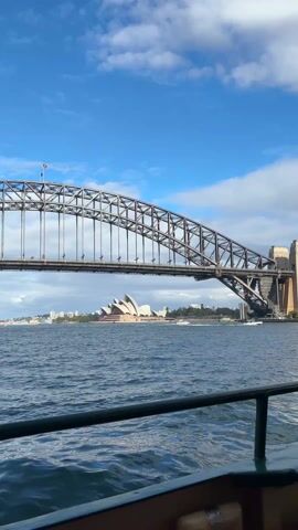 The video shows the Sydney Opera House and the Sydney Harbour Bridge from the vantage point of a boat. The Opera House is in the distance, distinct with its white, shell-like structure. The bridge looms overhead, its metal structure dominating the frame. The water is choppy, reflecting the blue sky with scattered white clouds. The boat's railing is visible in the foreground.
