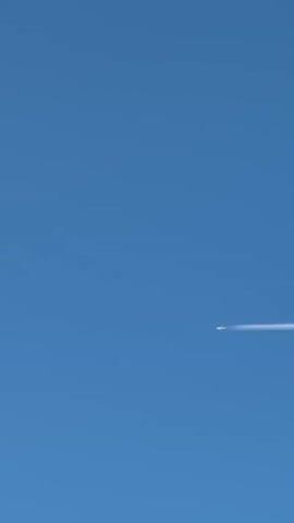 The video shows a clear blue sky. A single airplane is visible as a small white dot in the right portion of the frame. A long white contrail extends from the back of the airplane, stretching horizontally across the sky to the right. The airplane is moving from left to right, leaving the contrail behind. The sky is completely cloudless.