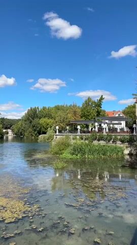 The video opens with a view of a calm river on a sunny day. The water is somewhat murky with green algae floating on the surface and obscuring the riverbed. On the right bank, there is a building with an outdoor seating area and vegetation along the water's edge. In the background, trees form a lush green backdrop against a bright blue sky with scattered white clouds. The camera then pans to the left, revealing more of the river and the trees on the opposite bank. Further along the riverbank, there are more buildings, including one with a distinctive red brick chimney. The camera continues to pan, showing houses and trees lining the river. The video concludes with a shot of a stone bridge with arched spans, supported by pillars. The bridge is adorned with decorative lamps and planters. The sky above the bridge is blue with white clouds.