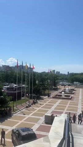The video shows a view from an elevated position looking down on a plaza on a sunny day. The plaza has a patterned tiled ground with square sections of light and dark tiles. In the background, there is a row of flags on poles and trees. Further in the distance, there are buildings and a clear blue sky. Several people are scattered throughout the plaza, walking or sitting on benches. There are also some cars parked on the left side of the plaza. In the foreground, there is a large concrete structure and some stairs. There is a vehicle parked on the lower left. The video captures a serene outdoor scene with people enjoying the open space.