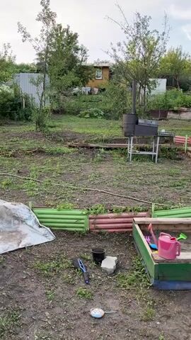 The video shows a panoramic view of a backyard with various elements. In the foreground, there's a sandbox with toys, including a pink watering can, a yellow duck toy, and a blue shovel. To the left of the sandbox is a black bucket and a gray brick. Further back, there's a metal barbecue grill with a chimney. A wooden swing set is visible, with what appears to be cucumbers growing on it. A large bush with white flowers is situated next to the swing set, and several potted flowers are nearby. A house with a dark roof is in the background, along with trees and other vegetation. A dark-colored SUV is parked on the right side of the yard, next to a blue barrel and an orange watering can. The ground is mostly dirt and grass.