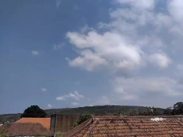 The video shows a view of the sky from a window, with clouds passing overhead. Below the sky, there are rooftops of houses and a distant view of a hill covered in trees. The overall scene is one of an outdoor environment, likely a residential area.