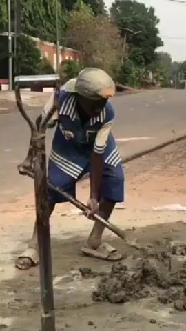 The video shows a young boy, likely of African descent, standing beside a pile of wet cement, holding a shovel. He is wearing a baseball cap, a blue and white patterned shirt, blue shorts, and sandals. He looks off-camera. To his left, an older-style, long-handled hand-operated cement mixer stands. He then seems to pause and another man, also of African descent, walks quickly past him in the background. The second man is wearing a blue t-shirt, appears older, and has cement dust on his clothing. The setting appears to be a street scene, possibly in a residential area, with buildings and trees visible.