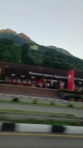 The video shows a panning shot of a street scene in a mountainous area. In the foreground, there is a road with white and black striped barriers. Beyond the road, there is a sidewalk with some greenery. Further back, there is a series of buildings with a distinct architectural style, featuring arched windows and balconies. Some of the buildings are tall and multi-story, while others are shorter. There are also flags or banners attached to some of the buildings. In the background, lush green mountains rise up, with a clear sky above. The lighting suggests it is either early morning or late afternoon. The scene appears to be a resort or a town located in a mountainous region. There are no people clearly visible in the video, and no specific actions or interactions are captured. The overall impression is a scenic view of a developed area nestled within a natural landscape.