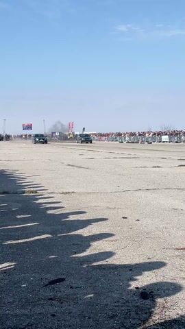 The video shows two cars, a dark grey Audi and a dark grey Volkswagen Golf, drag racing on a paved surface. They are in the middle of a wide track lined with spectators behind barriers on both sides. The Audi appears to be ahead of the Golf. Both cars are kicking up some dust as they accelerate. The day is sunny with a clear blue sky. The camera is positioned low to the ground, showing the shadows of people and objects in the foreground. After the cars pass the camera, it pans slightly to follow their movement as they continue down the track. The background is blurred with a banner and some green land visible in the distance.