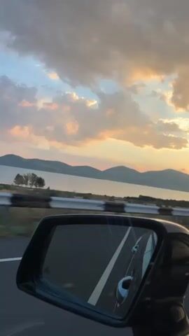 This is a vertical video taken from the passenger seat of a moving car. The scene captured in the video is a landscape view of a body of water and mountains under a cloudy sky at sunset. A side mirror of the car is visible in the foreground, reflecting the road and sky. Trees and vegetation line the side of the road. The sun is setting behind the mountains, casting a warm glow on the clouds.