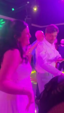 A group of people are dancing in a dimly lit room with red and blue spotlights. A woman in a white dress is dancing in the foreground, her hair is dark and wavy. A man in a white collared shirt and light-colored pants is dancing next to her. Other people are visible in the background, some dancing and some standing. The room appears to be a club or a party venue. The lighting is dynamic, shifting between red and blue hues.