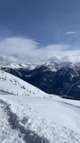 The video shows a mountain range covered in snow under a cloudy sky. The landscape is predominantly white with patches of dark forest and rock visible through the snow cover. The camera pans across the scene, showing the vastness of the mountain range and the valley below. The sun occasionally peeks through the clouds, casting a bright light on the snow-covered peaks. There are tracks in the snow, indicating that people have been skiing or snowboarding in the area. The video captures the serene and majestic beauty of a winter mountain landscape.
