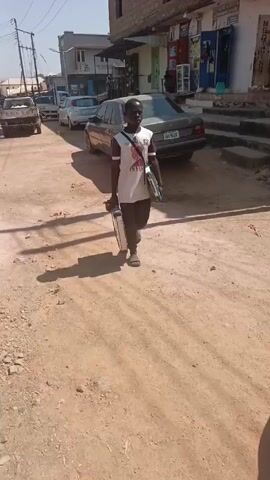 A man walks on a dirt road, carrying two metal briefcases in his left hand and a blue case slung over his shoulder. He wears a white t-shirt, dark pants, and sandals. The road has cars parked along it and a building with shops on the right side. The day appears sunny.