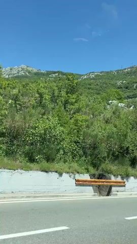 The video shows a moving perspective from a vehicle on a paved road. To the left, a steep, rocky hillside covered in green vegetation rises under a clear blue sky. The road has white lane markings. In the distance, on the hillside, there are some buildings and vehicles. The camera pans slightly to the right, revealing more of the lush green landscape and the expansive sky.