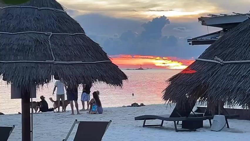 The scene is a beach at sunset, with a vibrant orange and red sky reflecting on the water. There is a small island with a structure on it in the distance. Two thatched umbrellas provide shade on the white sandy beach, with lounge chairs positioned beneath them. A group of people, including children and a dog, are gathered near the water's edge, appearing to observe the sunset.  One person is in the water. The scene is tranquil and serene.