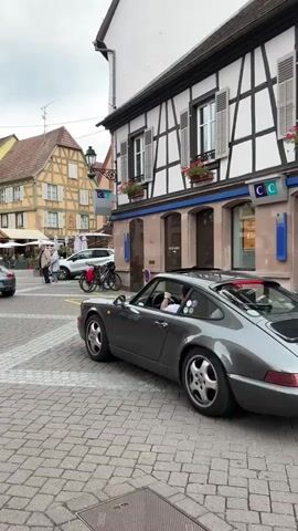 A gray Porsche 911 is seen turning in a cobblestone square in front of buildings with half-timbered facades. Several people are walking in the square, and other vehicles are parked in the background. The sky is overcast.