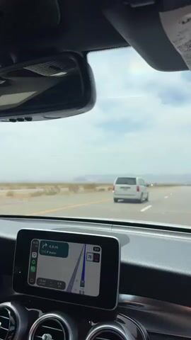 The video shows a view from inside a car traveling on a highway. The dashboard is visible in the foreground, with a navigation screen displaying a map. Outside the car, a white minivan is driving in front, in the same lane. The landscape appears to be a desert or arid region, with mountains visible in the distance under a hazy sky. The weather appears to be bright and possibly hot.